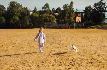 Photo for A seven-year-old girl with pigtails plays with a west highland white terrier on the beach near the lake. Lifestyle concept. - Royalty Free Image