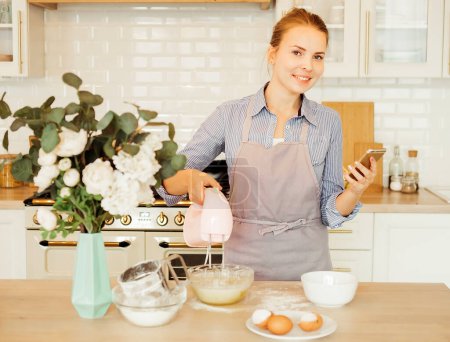 Téléchargez les photos : Young blonde woman uses mixer and whips cream for cake, stands in modern kitchen - en image libre de droit
