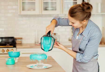 Foto de Young blonde woman decorating a cake with blue icing, modern kitchen, freelancing and hobby concept - Imagen libre de derechos
