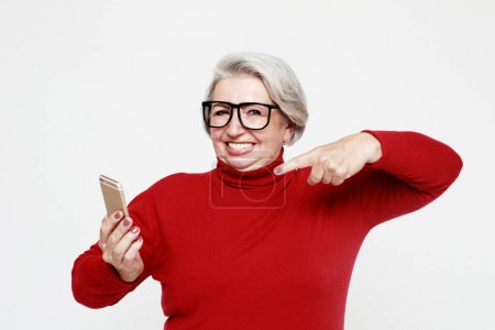 Téléchargez les photos : Lifestyle, technology and old people concept: A picture of mature woman wearing red sweater with new smartphone. She has tested it and admitted this phone is a good one. - en image libre de droit