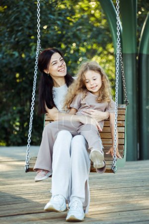 Photo for Mom and daughter swing on a swing. Caucasian young happy woman and her little girl have fun on the playground. - Royalty Free Image