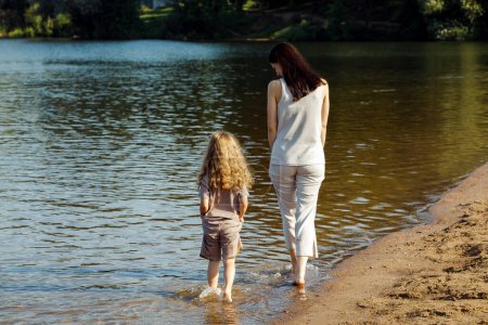 Téléchargez les photos : A five-year-old cheerful girl and her mother are walking by the lake, barefoot in the water, having fun on a summer day. Happy time. - en image libre de droit