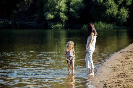 Foto de A five-year-old cheerful girl and her mother are walking by the lake, barefoot in the water, having fun on a summer day. Happy time. - Imagen libre de derechos