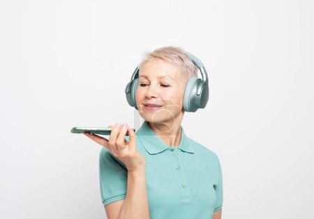Photo for Smiling mature woman in headphones with short hair recording voice message on phone close up, happy older female chatting online or speakerphone - Royalty Free Image