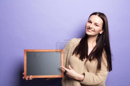 Téléchargez les photos : Young smiling brunette woman holding a chalkboard isolated on purple background. - en image libre de droit