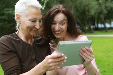 Téléchargez les photos : Two happy old women laugh merrily and look at the screens of tablet. Having fun in retirement on a summer afternoon. Golden age. - en image libre de droit