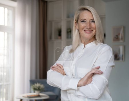 Photo for Smiling confident stylish mature middle aged woman standing at home office. Old senior businesswoman, 60s gray-haired lady executive business leader manager looking at camera arms crossed, portrait. - Royalty Free Image