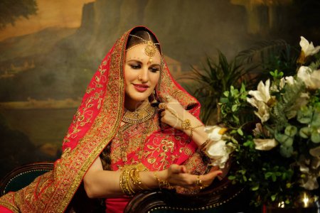 Foto de Charming Indian bride dressed in Hindu red traditional wedding clothes embroidered with gold and a veil smiles tender sitting on sofa near flowers. - Imagen libre de derechos