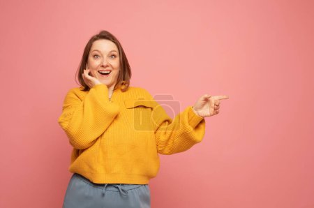 Photo for Surpeised young woman pointing with finger at blank copy space aside over pink background. - Royalty Free Image