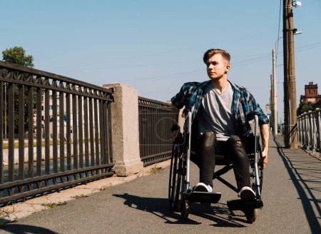 Foto de A young disabled man rides in a wheelchair across a bridge, the young male dressed in a plaid shirt and jeans. Lifestyle concept. - Imagen libre de derechos
