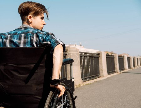 Photo for A young disabled man rides in a wheelchair across a bridge, the young male dressed in a plaid shirt and jeans. View from the back. - Royalty Free Image
