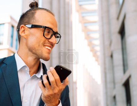 Téléchargez les photos : Handsome young man wearing eyewear recording audio message on smartphone while walking on city street. Man use smartphone to send voice messages outdoors. - en image libre de droit