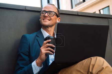 Foto de Joven hombre elegante que trabaja en el ordenador portátil mientras está sentado en el banco al aire libre y beber café. Negocios, gente, tehnología y concepto de estilo de vida. - Imagen libre de derechos