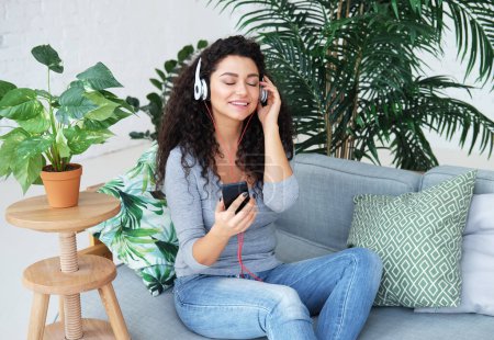 Foto de Young beautiful african american woman with long hair relaxing on sofa and listening to music using headphones. Lifestyle concept. - Imagen libre de derechos