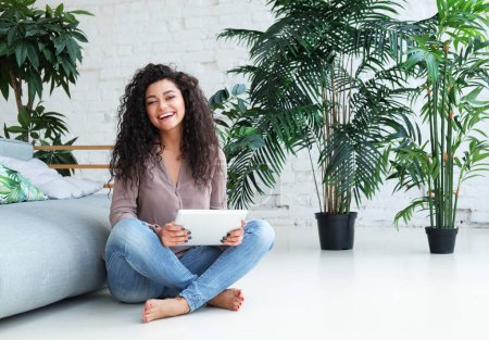 Photo for Beautiful Afro American female is using a digital tablet while sitting on the floor near sofa at home. Lifestyle concept. - Royalty Free Image