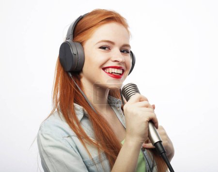 Téléchargez les photos : Cheerful young red-haired woman wearing headphones holding a microphone and singing. Lifestyle concept. - en image libre de droit