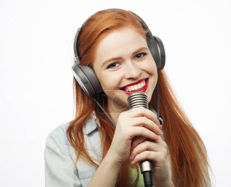 Téléchargez les photos : Cheerful young red-haired woman wearing headphones holding a microphone and singing. Lifestyle concept. - en image libre de droit