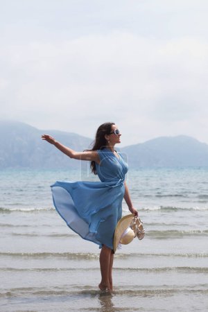 Foto de Young brunette woman in a blue dress walking barefoot on a beach and dangles his feet in the water. Lifestyle concept. - Imagen libre de derechos