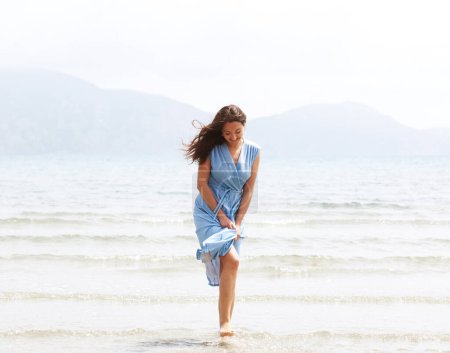 Photo for Young brunette woman in a blue dress walking barefoot on a beach and dangles his feet in the water. Lifestyle concept. - Royalty Free Image