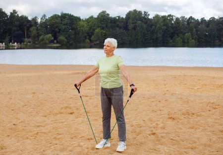 Téléchargez les photos : Elderly woman wear sportswear doing exercises outdoor on the beach in the morning using resistance rubber bands. Healthy lifestyle, active retired life and sporty time. - en image libre de droit