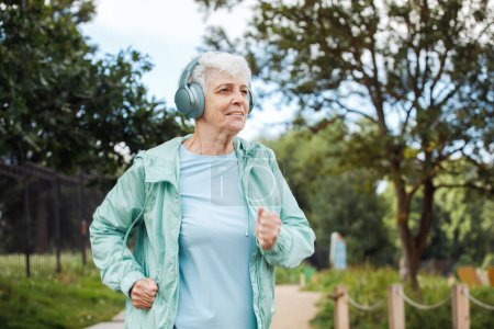 Photo for An elderly woman with a short haircut dressed in sportswear is jogging in the park while listening to music. Lifestyle and old people concept. - Royalty Free Image