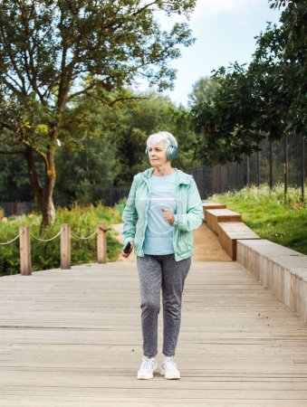 Foto de An elderly woman with a short haircut dressed in sportswear is jogging in the park while listening to music. Lifestyle and old people concept. - Imagen libre de derechos