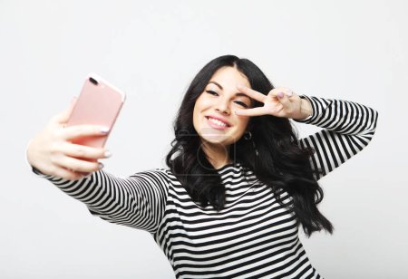 Téléchargez les photos : Fashion curvy brunette woman wearing striped sweater taking photo makes self portrait on smartphone over white background - en image libre de droit