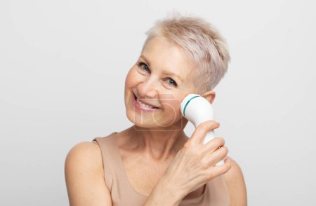 Foto de Beautiful 60s woman with short hair holding a massager in her hand and use on face. Spa technique skincare ads. - Imagen libre de derechos