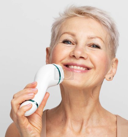 Photo for Beautiful 60s woman with short hair holding a massager in her hand and use on face. Spa technique skincare ads. - Royalty Free Image