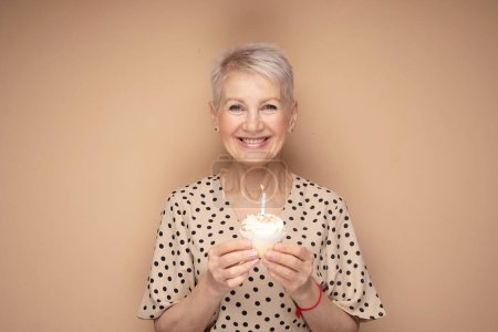 Photo for Cheerful elderly woman with short haircut holds a cupcake with a candle in her hands, celebrates a birthday and laughs - Royalty Free Image