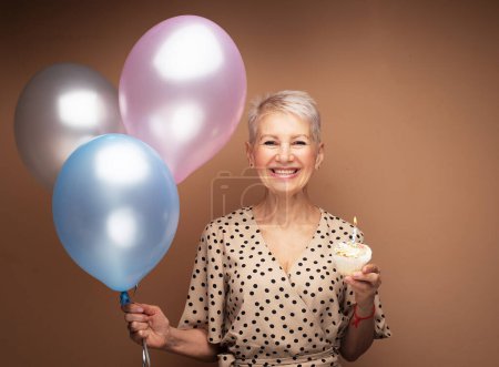 Photo for Beauitful happy elderly female with cupcake with candle and balloons in her hands. Portrait over beige background. - Royalty Free Image