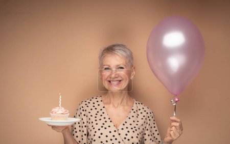 Téléchargez les photos : Belle femme âgée heureuse avec cupcake avec bougie et ballons dans ses mains. Portrait sur fond beige. - en image libre de droit