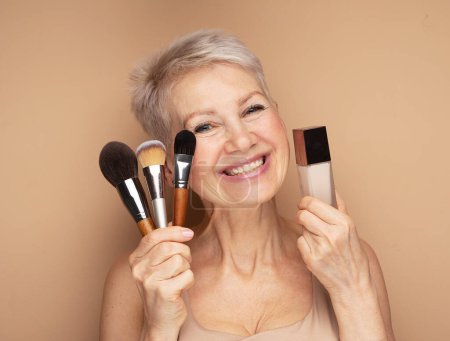 Photo for Charming elderly woman holds makeup brushes and foundation in her hands over beige background - Royalty Free Image