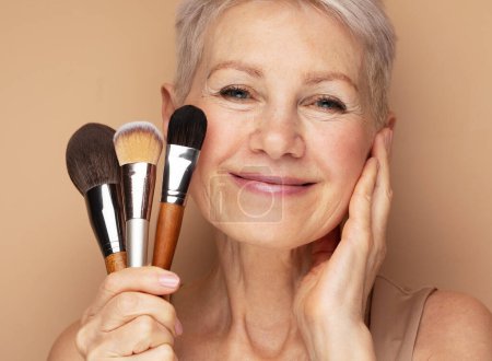 Photo for Charming elderly woman holds makeup brushes over beige background - Royalty Free Image