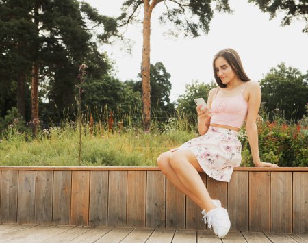 Photo for Young smiling woman in casual wear sitting on bench and use mobile cell phone chat online in social network. Summer park. Lifestyle concept. - Royalty Free Image
