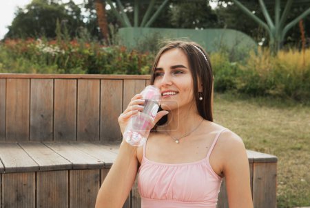 Photo for Beautiful smiling thirsty woman sitting on bench and drinking fresh bottled water in summer park. - Royalty Free Image