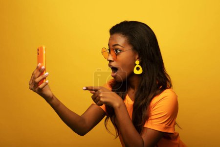 Photo for Lifestyle, tehnology and people concept: Portrait of inspired dreamy american young surprised woman use her smartphone over yellow color background - Royalty Free Image