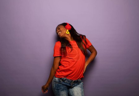 Téléchargez les photos : Enthousiaste mélange race femme avec les cheveux longs écouter de la musique préférée par écouteurs sur fond violet - en image libre de droit