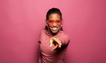 Téléchargez les photos : Belle jeune femme afro porter des vêtements décontractés et des lunettes de soleil danse recherche vide espace isolé couleur rose fond - en image libre de droit