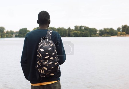 Foto de Un joven negro en ropa deportiva con una mochila camina cerca del lago. Día de verano. Vista trasera. - Imagen libre de derechos