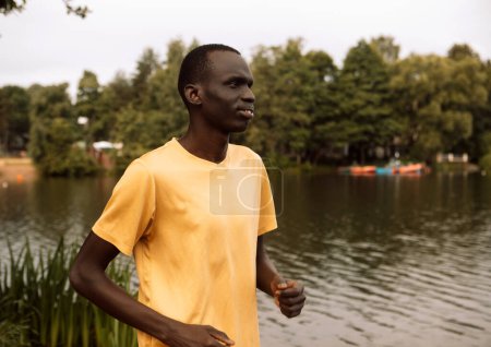 Foto de A young black man in a yellow T-shirt is jogging near a lake. Morning. Summer day. Lifestyle and sport concept. - Imagen libre de derechos
