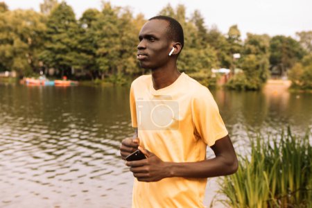 Foto de A young black man in a yellow T-shirt is jogging near a lake. Morning. Summer day. Lifestyle and sport concept. - Imagen libre de derechos