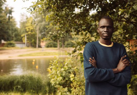 Téléchargez les photos : Portrait of a young african man who stands with his arms crossed on a summer day in the park - en image libre de droit