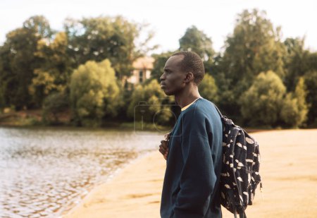 Foto de Un joven negro en ropa deportiva con una mochila camina cerca del lago. Día de verano. - Imagen libre de derechos
