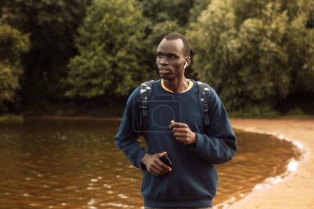 Foto de A young black man wearing casual jogging near a lake with headohones. Morning. Summer day. Lifestyle and sport concept. - Imagen libre de derechos