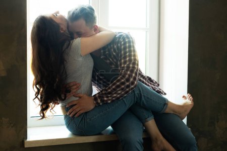 Foto de Pareja joven abrazándose y besándose mientras se sienta en el alféizar de la ventana. Feliz vida familiar. Amor.. - Imagen libre de derechos