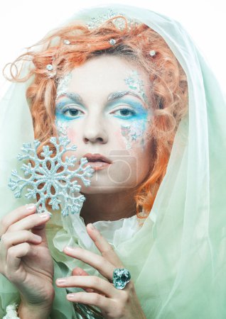 Foto de Concepto de fiesta, navidad y gente: Una joven hermosa pelirroja con un maquillaje creativo brillante en un estilo de Año Nuevo sostiene un copo de nieve plateado cerca de su cara. Reina de la nieve, hada. - Imagen libre de derechos