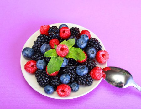 Photo for Various berries in a white plate. Blueberries, blackberries and raspberries are delicious diet berries. Plate with spoon on pink background. - Royalty Free Image