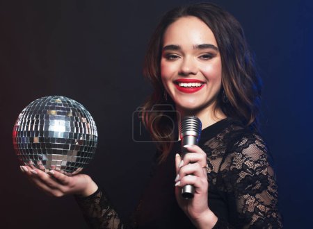 Téléchargez les photos : Mode de vie, fête et concept de personnes : jeune femme portant une robe noire, tenant le ballon disco et chantant dans le microphone sur fond sombre - en image libre de droit