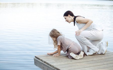 Foto de Una joven madre y su hija de cinco años observan los peces en el lago, de pie en el puente. Feliz verano. El concepto de conocimiento del mundo y de la familia. - Imagen libre de derechos